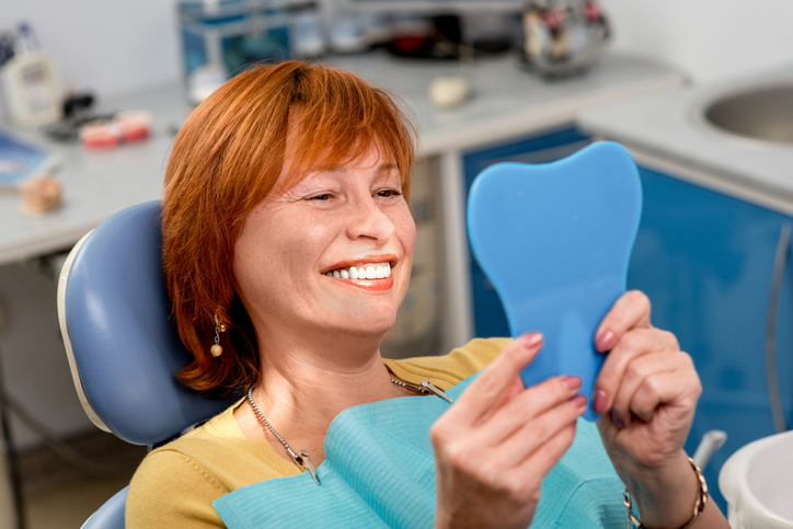 Smiling woman with new dental implants sitting in the dental office and looking at the mirror
